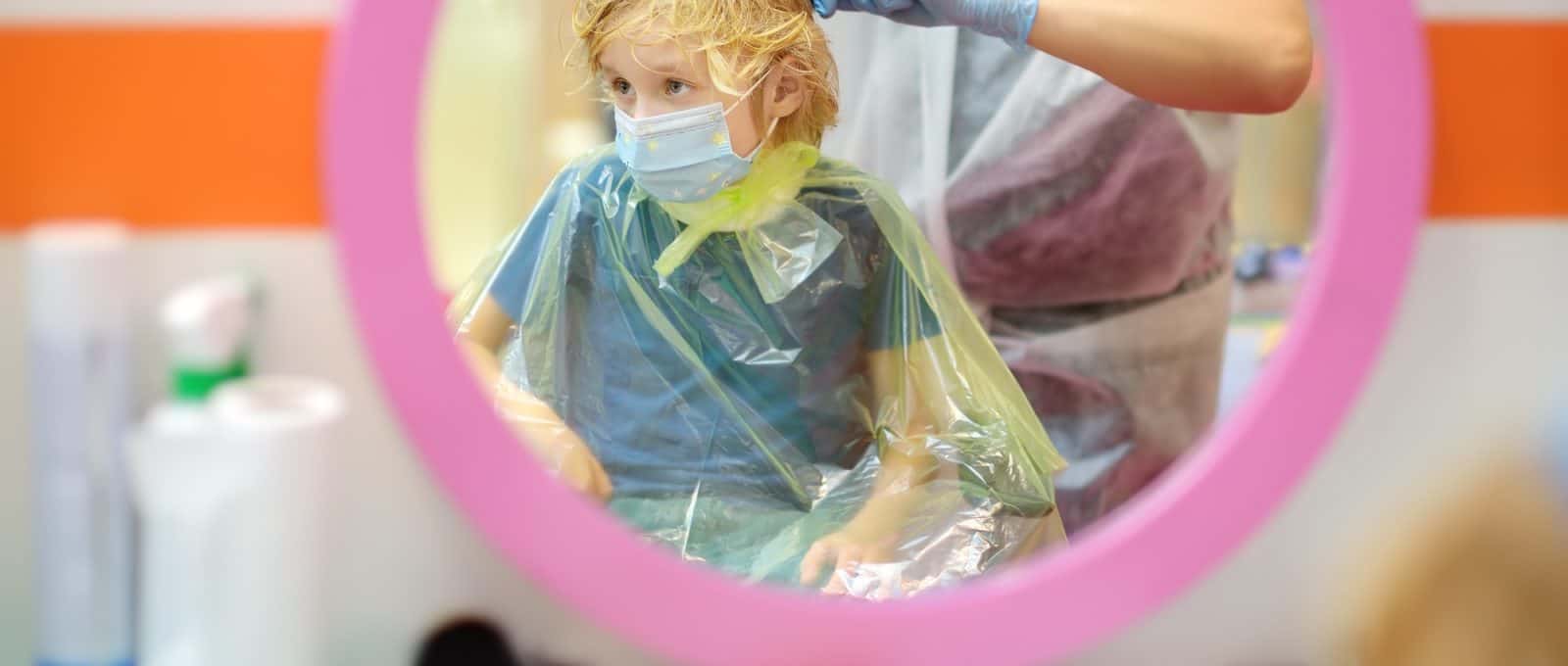 Preschooler boy wearing face mask getting haircut in salon during coronavirus epidemic.